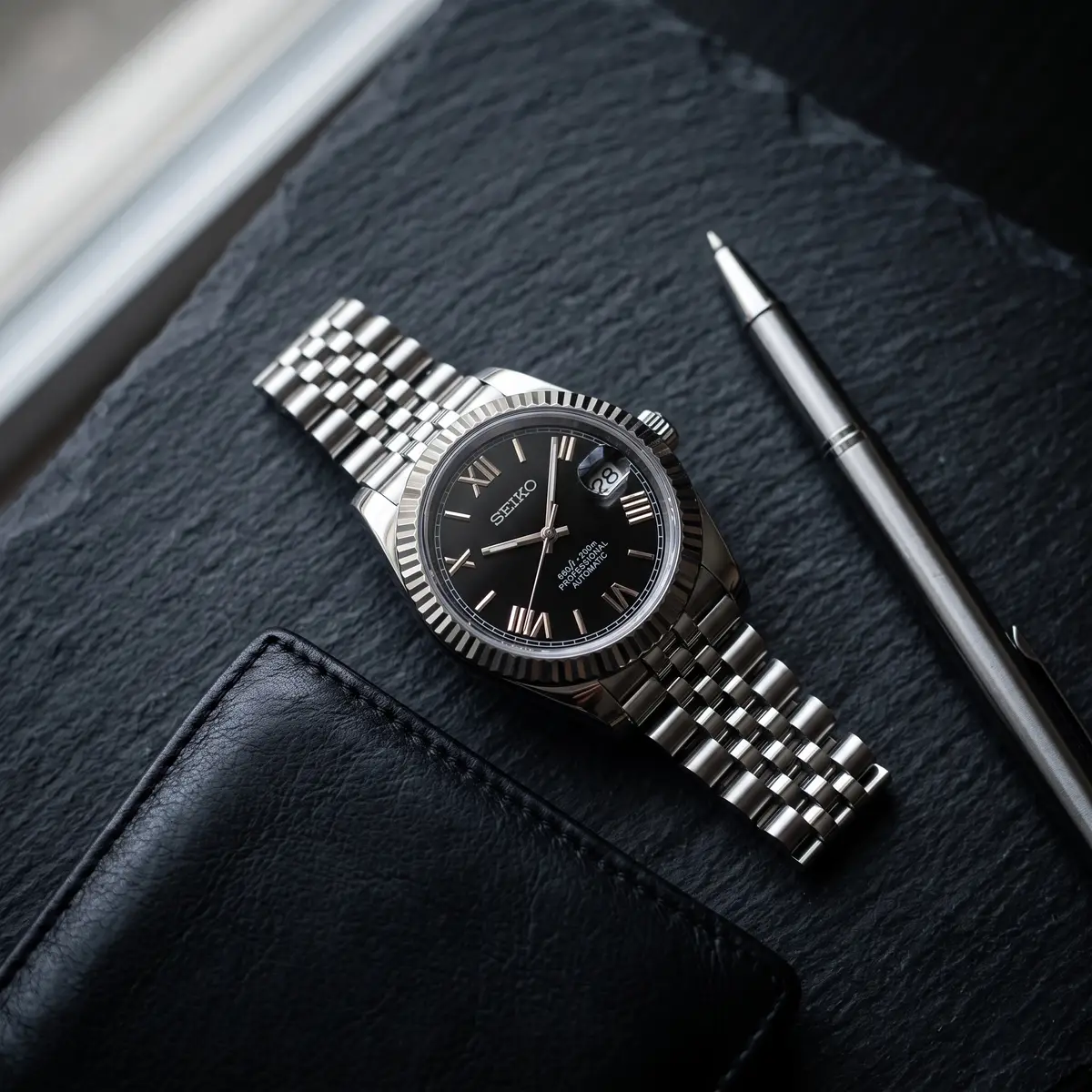 Seiko Mod Uhr DATE MOD | Stahl, schwarzes Zifferblatt mit römischen Ziffern – Automatik – Flatlay auf Schiefer – MedoMods